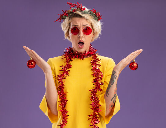 周围笨手笨脚的金发女郎头戴圣诞花环 脖子上戴着金箔花环 手里拿着圣诞饰品 看起来在紫色的墙上与世隔绝头金发女人
