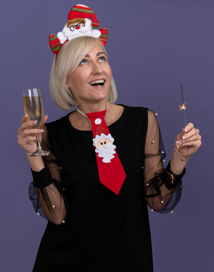 闪闪发光快乐的中年金发女人戴着圣诞老人的头带 打着领带 手里拿着节日火花和一杯香槟 孤零零地看着紫色的墙上头带中年领带