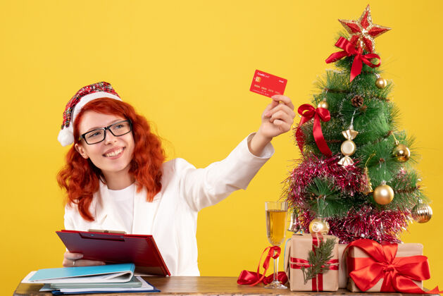 背景正面图：女医生坐在桌子后面 拿着黄色背景的银行卡 还有圣诞树和礼品盒生活人桌子