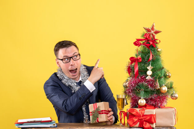 指点前视图中的快乐男子手指的东西坐在圣诞树附近的桌子上 黄色的礼物树坐着黄色