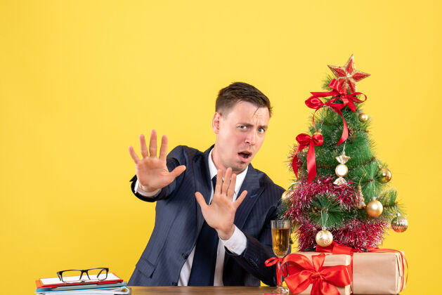 职业害怕的男人拒绝东西坐在圣诞树附近的桌子前 黄色的礼物西装新郎东西