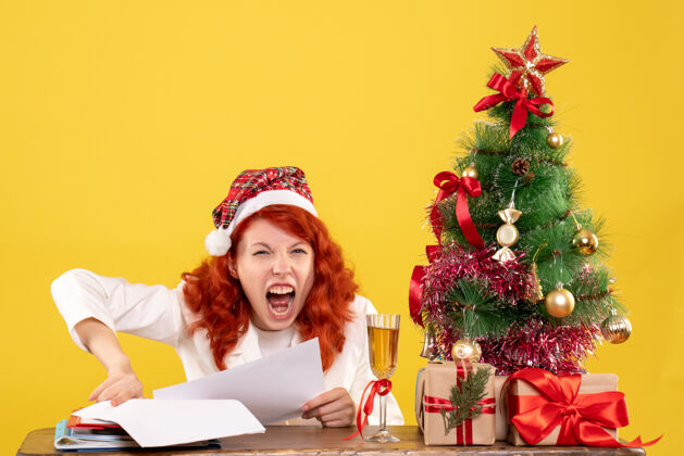 人前视图：女医生坐在桌子后面 拿着礼物 尖叫着放在黄色背景上 还有圣诞树和礼品盒礼物情感漂亮