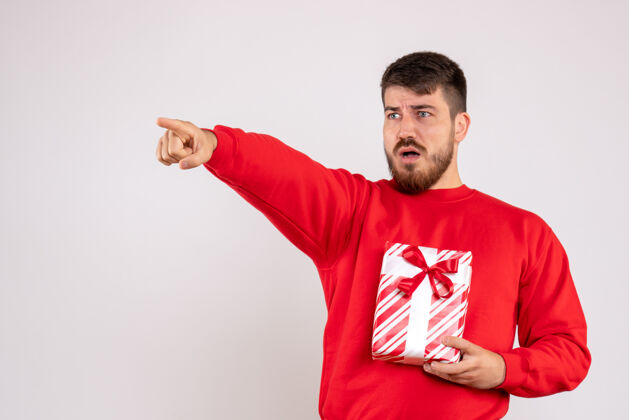 情感身穿红衫的年轻人拿着圣诞礼物指着白墙上的正视图成人球衣节日