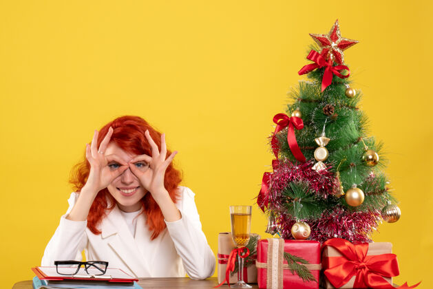医生正面图：女医生坐在桌子后面 拿着圣诞礼物 黄色背景上的树圣诞树礼物新郎