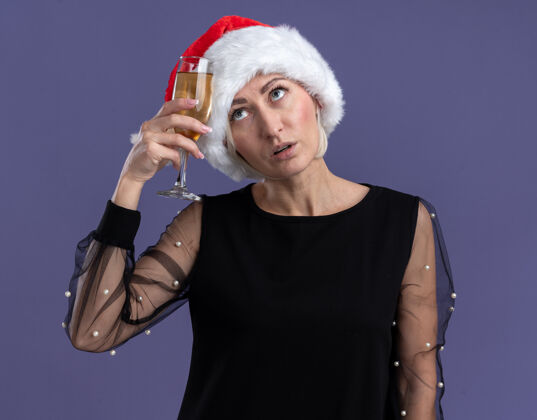 感动困惑的中年金发女人戴着圣诞帽抬头抚摸着头 紫色背景上隔着一杯香槟香槟圣诞快乐圣诞