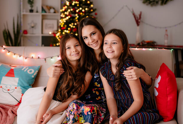 时间圣诞快乐的两姐妹和年幼的妈妈在家坐在客厅的沙发上 妈妈抱着女儿 大女儿看着身边的小女儿 妈妈看着相机家生活女儿