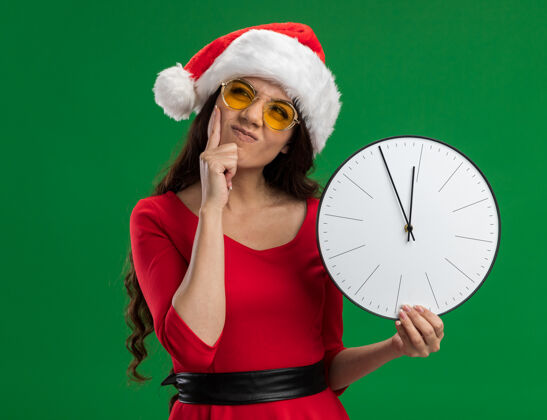女孩迷茫的年轻漂亮女孩戴着圣诞帽戴着眼镜拿着时钟抬头看保持手在下巴隔离在绿色背景上手年轻保持