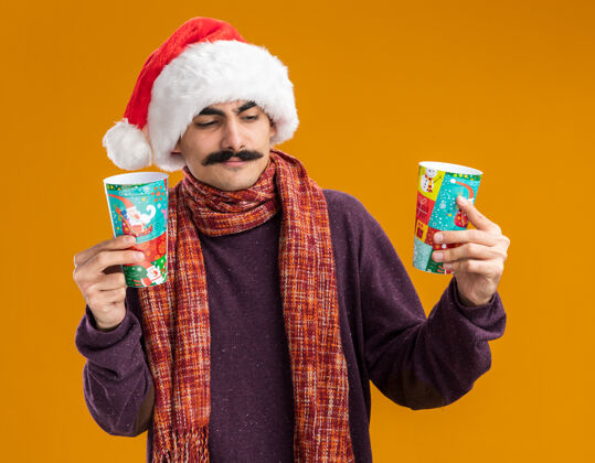 帽子一个留着胡子的男人戴着圣诞老人帽 脖子上围着暖和的围巾 手里拿着五颜六色的纸杯 站在橙色的背景上 看起来很困惑 试图做出选择杯子拿着选择
