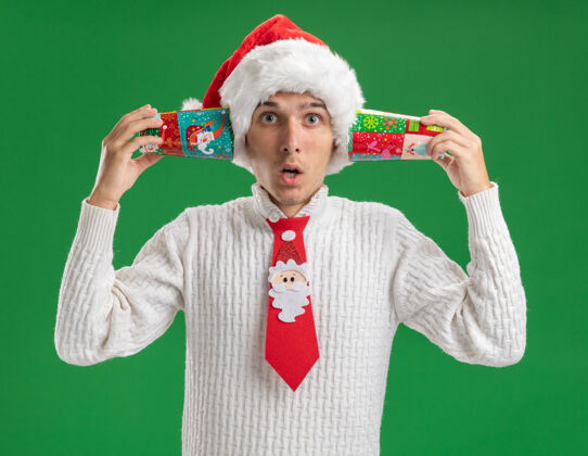 圣诞节印象深刻的年轻帅哥戴着圣诞帽 打着圣诞老人的领带 拿着塑料圣诞杯 耳边隔着绿色的墙壁塑料领带穿着