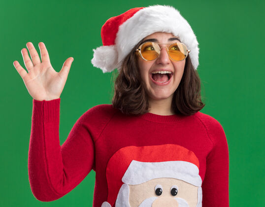 帽子穿着圣诞毛衣 戴圣诞帽 戴圣诞眼镜的快乐小女孩站在绿色的墙上 微笑着挥手致意毛衣高兴地站在一边
