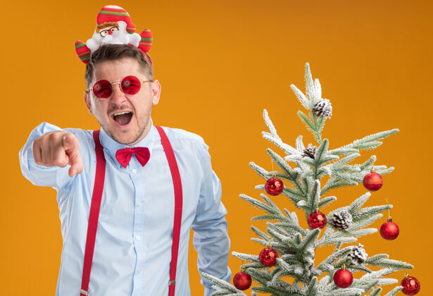 站着戴吊带领结的年轻人站在圣诞树旁 戴着圣诞老人和红眼镜 笑着用食指指着橙色的墙上的你们弓轮辋圣诞节