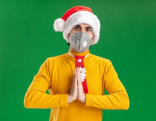 男人穿着黄色高领毛衣 戴着圣诞老人帽 打着滑稽的领带 戴着护面面具 手牵着手 严肃的脸站在绿色的墙上面部严肃领带