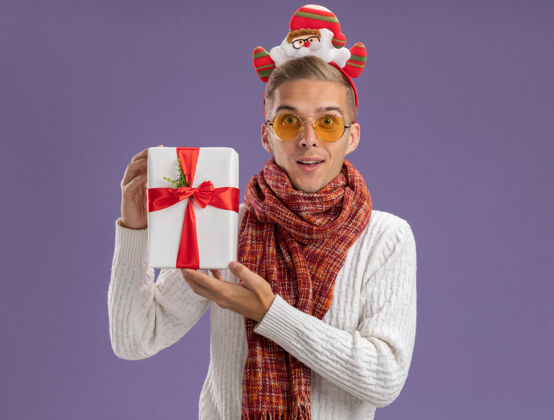 头带印象深刻的年轻帅哥戴着圣诞老人的头带和围巾拿着礼包隔离在紫色的墙上 有复制空间圣诞节帅哥圣诞老人