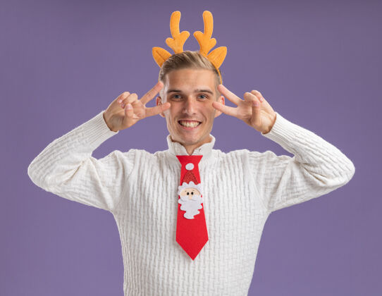 帅哥快乐的年轻帅哥戴着驯鹿鹿角的头带和圣诞老人的领带 在紫色的墙上 眼睛附近显示着v符号快乐年轻圣诞节