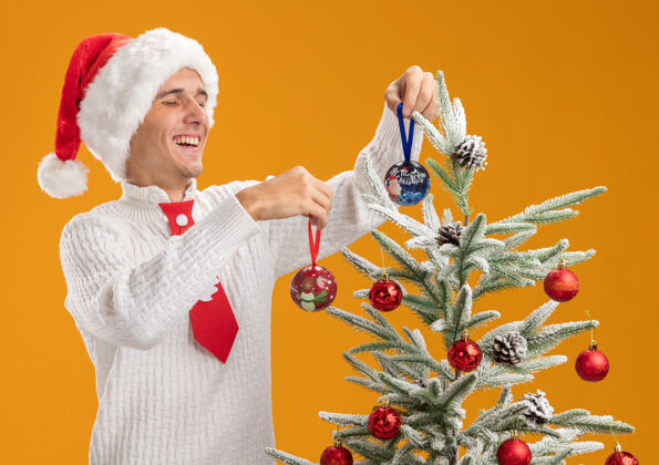 快乐快乐的年轻帅哥戴着圣诞帽 打着圣诞老人的领带 站在圣诞树旁 用橙色墙上的圣诞球装饰圣诞树年轻圣诞老人圣诞老人