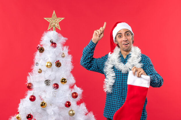情感情绪激动的年轻人戴着圣诞老人的帽子 穿着一件蓝色的衬衫 手里拿着圣诞礼物指着上面衬衫手指庆祝