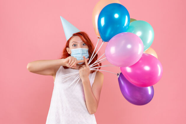 快乐前视图中的年轻女子拿着五颜六色的气球在无菌面具上粉红色的墙壁粉色气球年轻女性