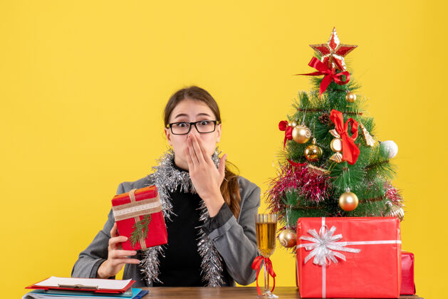坐前视图惊讶的女孩戴着眼镜坐在桌旁 把手放在嘴边圣诞树和礼物鸡尾酒女孩导演手