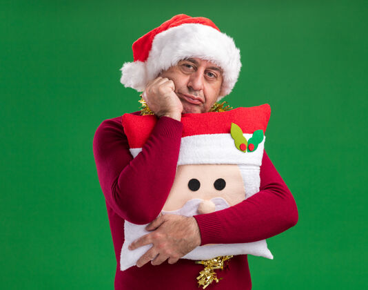 站着无聊的中年男子戴着圣诞老人帽 脖子上戴着金属丝 手拿圣诞枕头 头靠在手上看着相机 等待着绿色背景上的东西等待枕头金属片