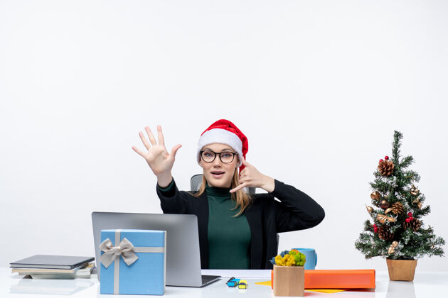 树戴着圣诞老人帽的年轻女子坐在桌子旁 手里拿着圣诞树和礼物电话工作场所制作