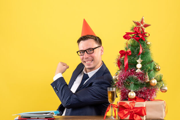 西装正面图：戴着派对帽的快乐商人坐在圣诞树旁的桌子旁 黄色的礼物聚会男人生意