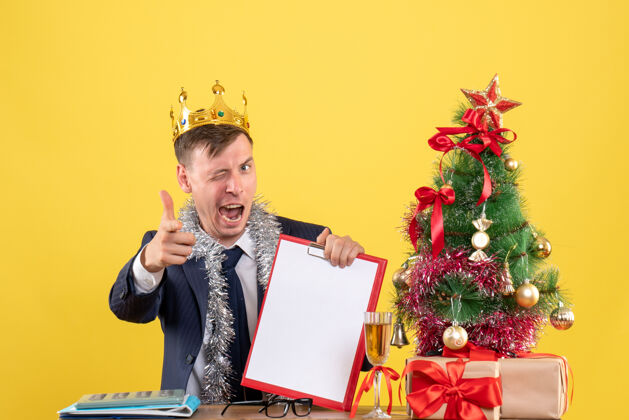 快乐商人眨着眼睛坐在圣诞树旁的桌子前 黄色的墙上摆着礼物微笑生意人礼物