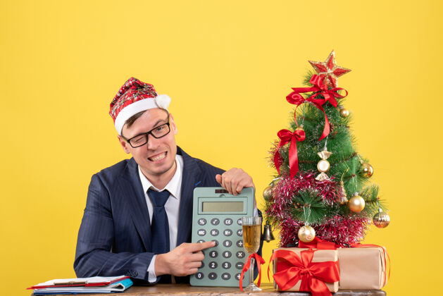 圣诞节前视图的商人显示计算器坐在圣诞树附近的桌子上 黄色的礼物商人专业礼物