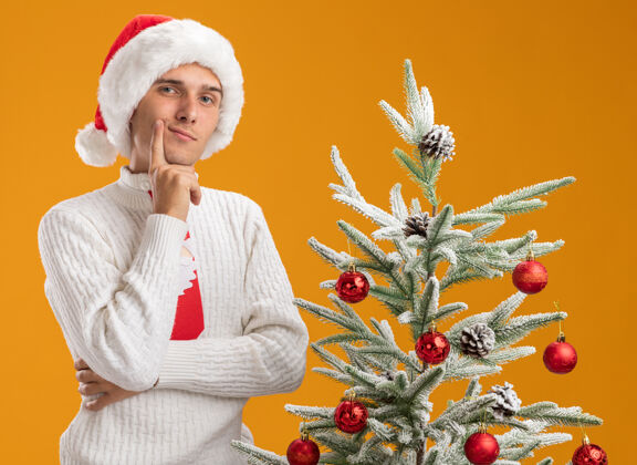 手体贴的年轻帅哥戴着圣诞帽 打着圣诞老人的领带 站在装饰好的圣诞树旁 手放在下巴上 看着橙色背景上孤立的摄像机装饰放帅气