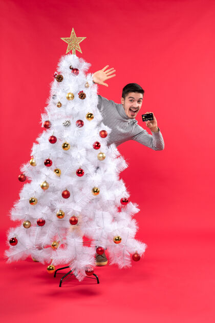 树快乐的年轻人站在装饰过的圣诞树后面 手里拿着红色的电话站立雪年轻人