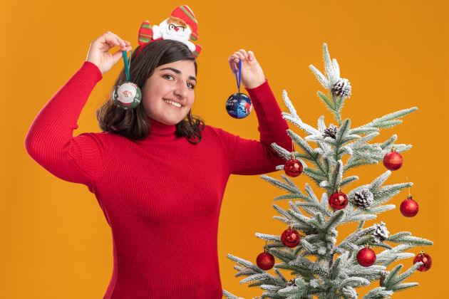 圣诞节穿着圣诞毛衣的快乐小女孩戴着滑稽的头带 手拿圣诞球 脸上带着微笑 站在橙色墙上的圣诞树旁头带微笑舞会