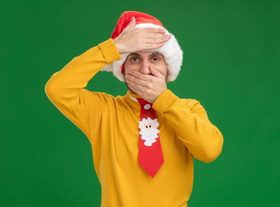 手戴着圣诞帽打着领带的年轻白种人看着相机 手放在嘴上和额头上 隔离在绿色背景上额头年轻圣诞节