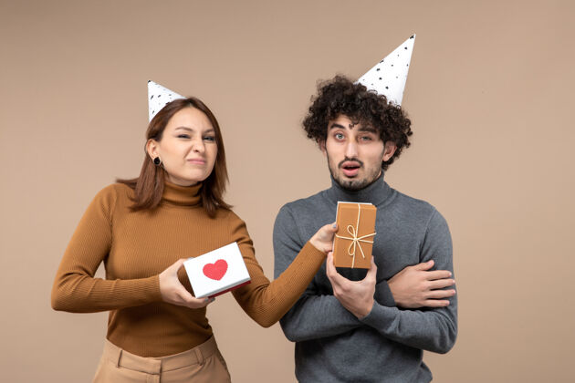 女人新年拍摄与年轻夫妇戴新年帽酸溜溜的脸女孩的心和困惑的家伙的礼物情侣帽子年轻夫妇