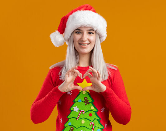 做身穿圣诞毛衣 戴圣诞帽的金发女郎面带微笑地看着摄像机 用手指在橙色背景上做着心形手势手指心年轻