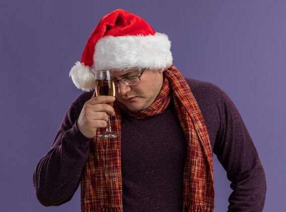 周围戴着眼镜 戴着圣诞老人帽子 脖子上围着围巾的疲惫的成年男子用一杯香槟酒触摸额头 闭着眼睛隔离在紫色背景上触摸围巾眼镜
