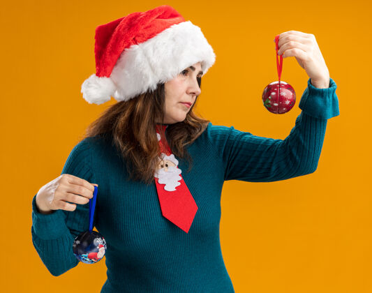 自信自信的成年白人妇女 戴着圣诞帽 打着圣诞领带 看着橙色背景上的玻璃球饰品 还有复制空间橙色圣诞老人球