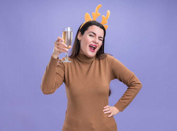 穿快乐的年轻漂亮女孩 穿着棕色毛衣 戴着圣诞发箍 手里拿着一杯香槟 手放在屁股上 隔离在蓝色的墙上毛衣圣诞节香槟