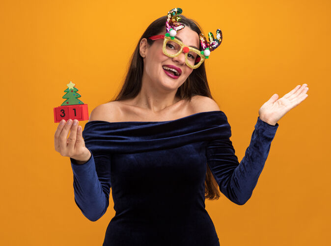 手微笑着的年轻漂亮的女孩穿着蓝色的裙子 戴着圣诞眼镜 手里拿着玩具 手被隔离在橙色的背景上女孩衣服微笑