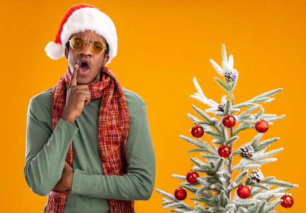 男人一个戴着圣诞帽 脖子上围着围巾的非裔美国人站在橙色墙上的圣诞树旁 惊讶不已惊讶站着到处都是