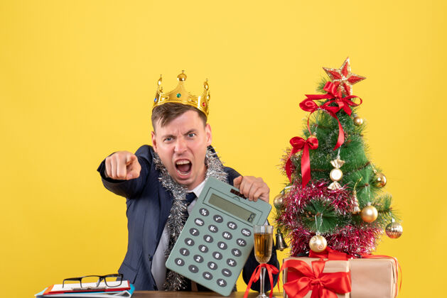 键盘前视图愤怒的男子指着摄像机坐在圣诞树附近的桌子和黄色墙上的礼物礼物键盘乐器前面
