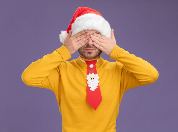 掩护年轻的白人男子戴着圣诞帽 打着领带 双手孤立在紫色的背景上遮住眼睛手帽子领带