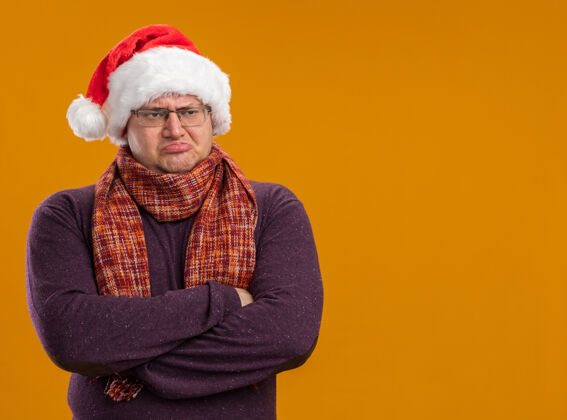 圣诞老人闷闷不乐的成年男子戴着眼镜 戴着圣诞帽 脖子上围着围巾 站在封闭的姿势 看着橙色背景上孤立的一面 留着复制空间站立封闭橙色