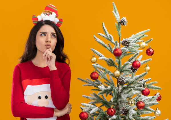 毛衣戴着圣诞老人头带和毛衣的可疑的年轻漂亮女孩站在装饰过的圣诞树旁 手放在下巴上 在橙色背景下孤立地抬头看橙色近树