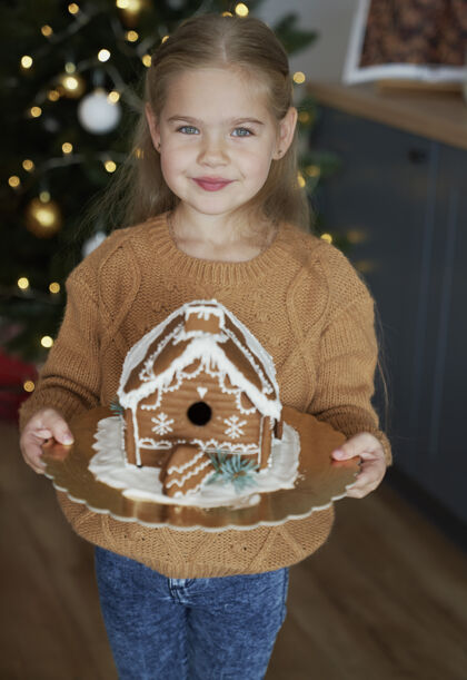 站立抱着装饰过的姜饼屋的小女孩季节乐趣圣诞树
