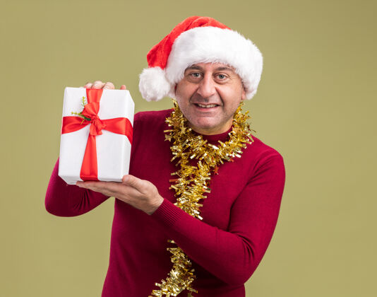脖子快乐的中年男人戴着圣诞帽 脖子上戴着金属丝 手里拿着圣诞礼物 脸上带着微笑 站在绿色的背景下看着相机圣诞老人礼物圣诞节