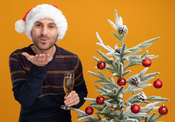 香槟高兴的年轻白人男子戴着圣诞帽 站在装饰好的圣诞树旁 拿着一杯香槟 看着摄像机 在橙色的背景下 孤零零地送上一吻看玻璃相机