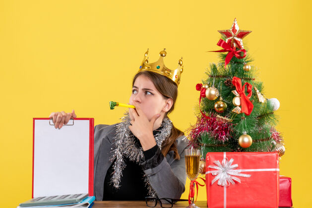 冬天正面图：戴着皇冠的年轻女孩坐在桌子旁 手里拿着圣诞树和鸡尾酒礼物文件年轻桌子