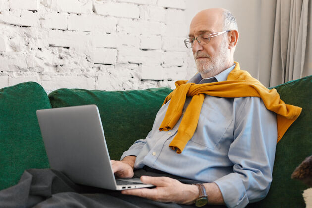 技术光头胡须的老年男性在家里用笔记本电脑连接无线高速互联网严肃专注的成熟商人在沙发上用笔记本电脑阅读商业新闻退休帅气电脑