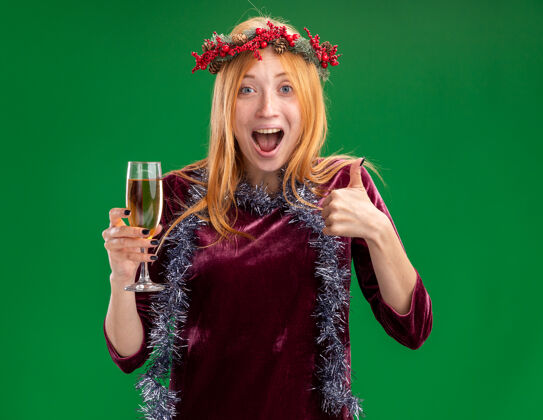 年轻兴奋的年轻漂亮女孩穿着红色的裙子 脖子上戴着花环和花环 手里拿着一杯香槟 在绿色的墙上孤立地竖起大拇指玻璃杯兴奋脖子