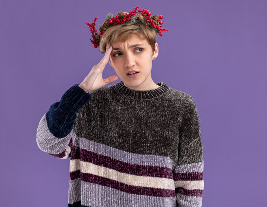 佩戴迷茫的年轻漂亮女孩戴着圣诞花环望着被隔离在紫色墙上的头圣诞节美丽花环