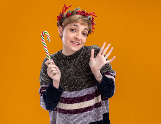 快乐快乐的年轻漂亮女孩戴着圣诞花环 手持圣诞甜手杖 孤零零地站在橙色的墙上穿着花环手杖